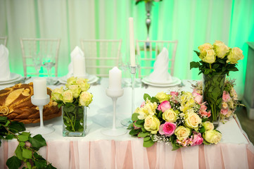 Wedding presidium table with pink tablecloth, floral arrangements: a bouquet of flower in a glass vase, candles in glass support, white curtains on background
