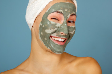 Beautiful cheerful teen girl applying facial clay mask. Beauty treatments, isolated on blue background.