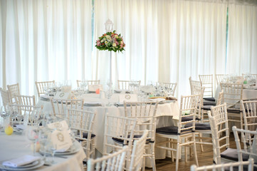 Lateral view of round wedding table with white tablecloth and all necessary supplies for dinner on them, bouquet of flowers; white curtains, beige floor, white chairs with blue pillows