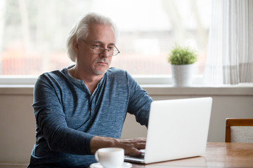 Serious aged man busy working at laptop drinking morning coffee at home, thoughtful senior male use...