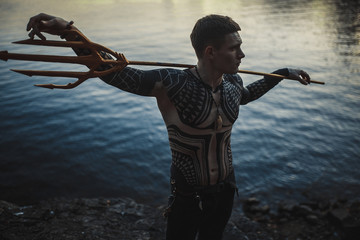 A young man with a trident in his shoulders against the background of water. - obrazy, fototapety, plakaty