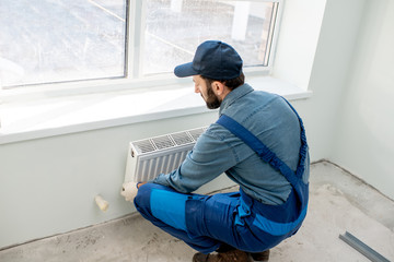 Workman mounting water heating radiator near the window in the white renovated living room