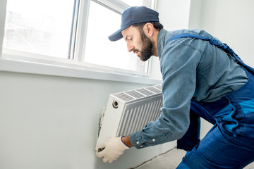 Workman mounting water heating radiator near the window in the white renovated living room - obrazy, fototapety, plakaty