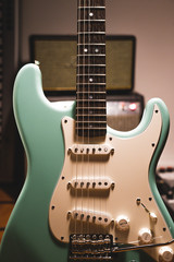 Fototapeta na wymiar Closeup of seafoam green custom electric guitar with wooden wenge fretboard and mother of pearl inlays, white pickup and pickguard in front of blurry background with amps music studio