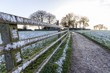 A light dusting of snow covers a field and fence along a curved path leading to a grove of bare trees and church in the background