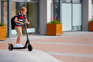 School boy in riding with his electric scooter in the city with backpack on sunny day. Child in colorful clothes biking on way to school