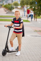 School boy in riding with his electric scooter in the city with backpack on sunny day. Child in...