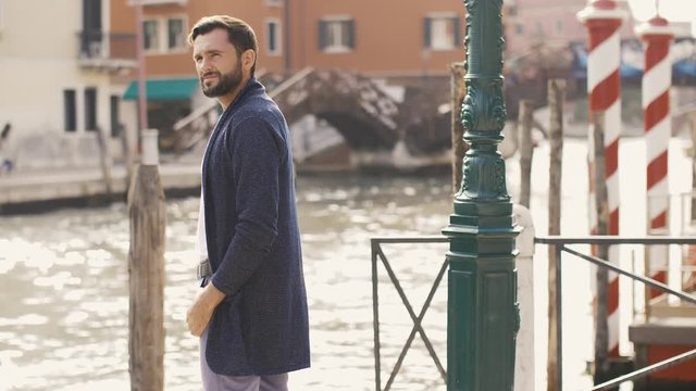 Handsome tourist man standing on the pier against beautiful view on venetian chanal with boats and gondolas in Venice, Italy. Beautiful slow motion 4k footage. Young Caucasian male in casual clothes