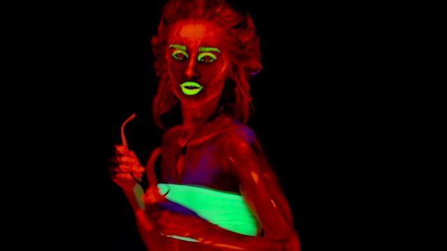 A beautiful young sexy half-naked girl putting fake glasses on and dancing with glowing paint on her body. Pretty woman with glowing bodyart in black lamp light.