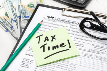 Sticker with phrase "TAX TIME", tax form, money, pencil, calculator and glasses onoffice table.