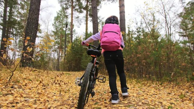 One caucasian children walk with bike in autumn park. Little girl walking black orange cycle in forest. Kid goes do bicycle sports. Biker motion ride with backpack and helmet. Mountain bike hardtail.