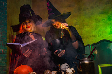 Portrait of smiling two witches in black hats reading book at table with pumpkin and skulls