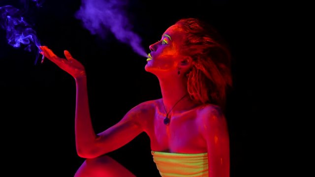 A beautiful young sexy girl with glowing paint on her body sitting and smoking a cigarette in black light. Pretty woman with glowing bodyart in black lamp light.