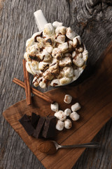 A mug of hot cocoa with toasted marshmallows next to a cutting board with chocolate chunks and cinnamon sticks, vertical