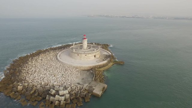 Amazing lighthouse in the middle of the ocean with view of Lisbon in the horizon. Aerial shot by drone in Portugal.