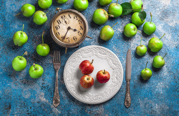 Organic apples arrangement overhead srved on rustic white plate and some spilled with fork and knife and old clock on painted blue background in studio