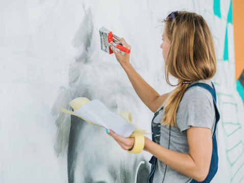 Beautiful Young Blonde Girl making graffiti of big eye with aerosol spray on urban street wall. Creative art. Talented student in denim overalls drawing picture