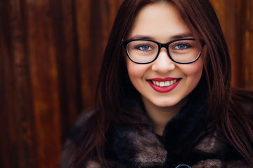 Close-up of a girl in glasses with a beautiful make-up in a fur coat that looks at the camera lens on the background of a wooden wall.
