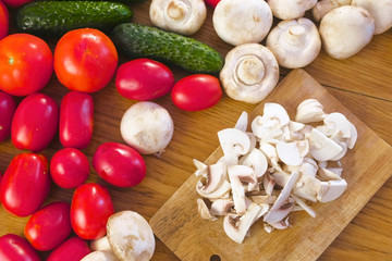 Cutting mushrooms champignons on wooden board on kitchen table with different vegetables.