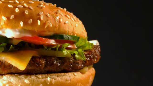 Juicy beef burger with cutlet, onion, vegetables, melted cheese, lettuce, sauce and topped sesame seeds. Isolated hamburger rotates on dark background, close-up view