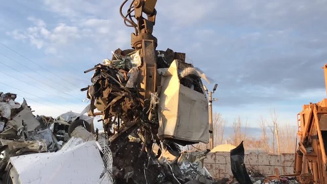 Powerful claw machine in junk yard grabs pile of metal into claws in slow motion before spinning in circles in sunlight thereafter. 