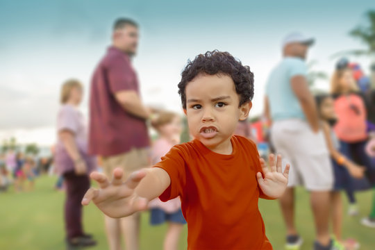 A small ethnic boy in a red shirt likes to show off while waiting in line. He pretends to be a super hero in front of the out of focus people in the background. 
