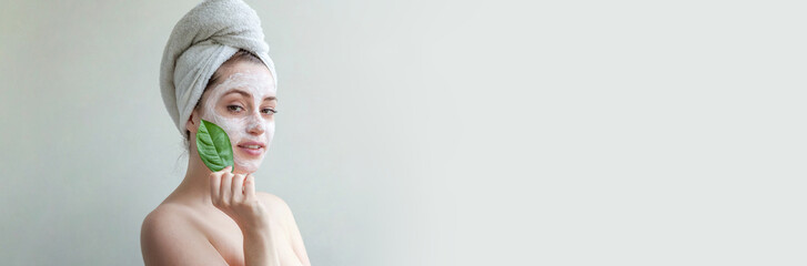 Beauty portrait of woman in towel on head with white nourishing mask or creme on face and green...
