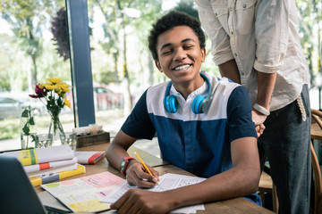 Smart student. Joyful Afro American man smiling to you while sitting at the table