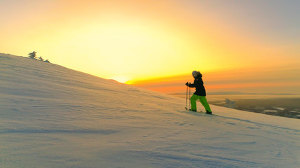Fototapeta na wymiar AERIAL: Active woman snowshoe trekking on snowy mountain slope at winter sunset