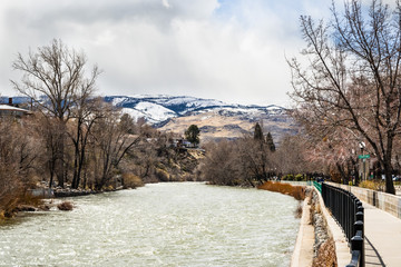 Truckee River flowing through downtown Reno on a cloudy spring day, Nevada; Paved walking path on the right; Sierra mountains covered in snow  in the background