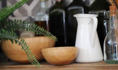 On the wooden kitchen table top there are wooden dishes. Green fern. White milk jug and bottles for oil and sauces.