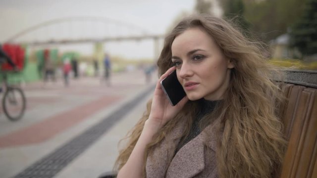 Medium close up beautiful young blonde woman with curly hair in trendy coat phone coversation sitting in the city park on bench on a warm day looking at the camera and smiling