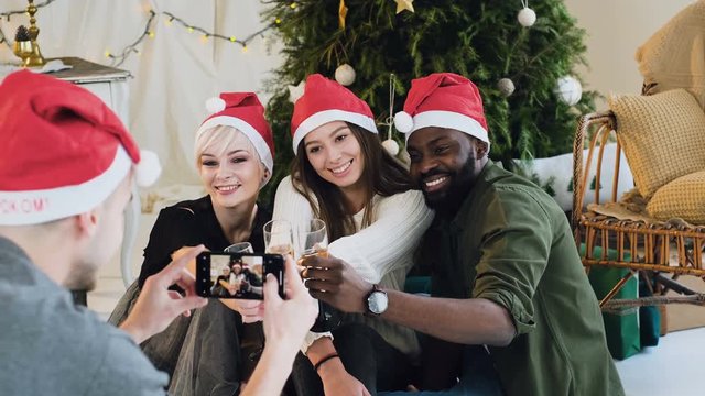 A group of people from different races posing and taking pictures on the phone. Cheerful friends in Santa hats make a photo on the smartphone at the christmas celebrate, good-looking young people