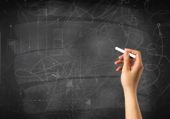 Female hand holding white chalk in front of a blackboard with scribbles and plans drawn on it 
