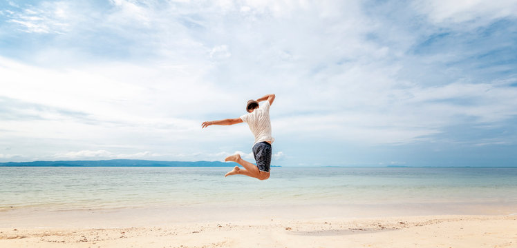 Young man in hat  jumping on the beach in front of ocean with feeling happy and freedom - Powered by Adobe