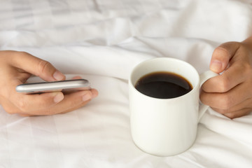 Coffee in bed; Hand holding a cup of black coffee and using mobile phone on bed. Lifestyle and simply perfect morning.