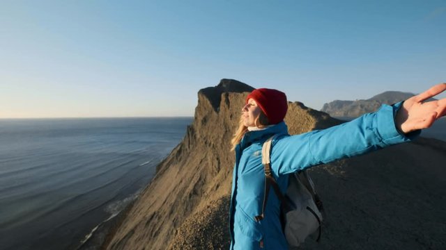 young attractive girl hiker goes along the sandy ledges or seaside cliff. Raises his hands up, looks at the golden sunrise or sunset. Enjoys a sense of success. Climbs on a high beautiful mountain.