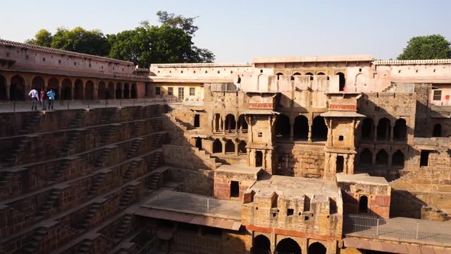 Panning shot of people viewing the beautiful architecture of the chand baori stepwell in Rajasthan. The geometrically made steps and stone palace served as a water storage for the dry areas around the