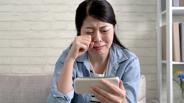 sad lady watching disaster movie on phone sitting on the couch in the living room at home with a bright background. depressed young woman with mobile cellphone crying. girl see sadness movie concept.