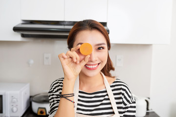 Cheerful woman is covering her eye with carrot in kitchen room at home