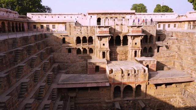 Panning shot of people viewing the beautiful architecture of the chand baori stepwell in Rajasthan. The geometrically made steps and stone palace served as a water storage for the dry areas around the