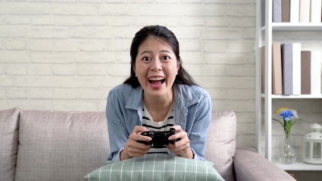Happy young girl self playing video games in cozy living room at home. lady with pillow on lap holding controller with an excited face. woman saying yes because of winning the game.