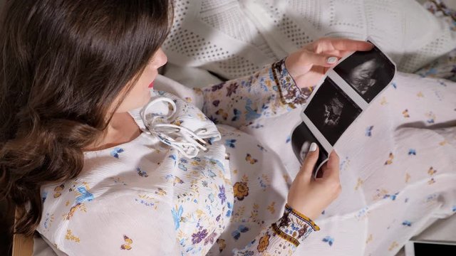 From above shot of pregnant woman holding fetal scan sitting on bed 