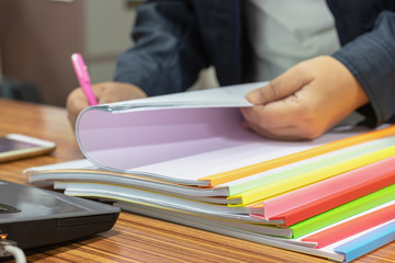 Teacher hand is holding pen for checking student homework assignments on desk in school. Unfinished paperwork stacked in archive with color plastic slide binder bars. Education and business concept.