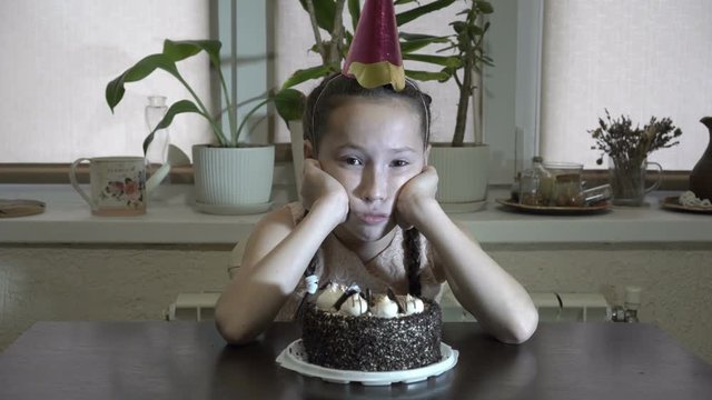 Sad little girl sitting at the table with his head propped up with his hands with a festive cap on his head and looks at the camera. Before her on the table is a cake. Close up. 4K. 25 fps.