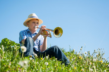 Boy in a straw hat sitting on a hill covered with green grass plays the trumpet.