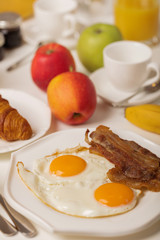 Breakfast time. Fried eggs and bacon. Croissants and orange juice, jam. Coffee with cream or milk. Fruits - bananas, red and green apples.