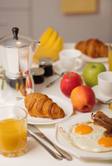 Breakfast time. Fried eggs and bacon. Croissants and orange juice, jam. Coffee with cream or milk. Fruits - bananas, red and green apples.