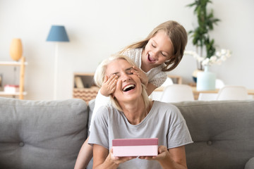 Little granddaughter closing eyes of excited old grandmother holding gift box, cute preschool child...