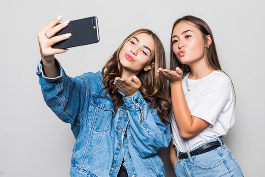 Two mixed race girls friends taking selfie with smartphone, blowing a kiss in the camera, isolated on white background